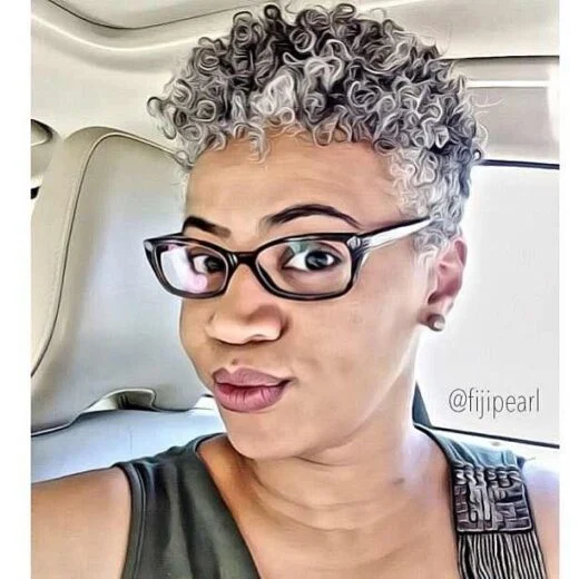 10 Stunning Pixie Haircuts for Black Women 10 A person with curly grey hair wearing glasses, seated in a car, dressed in a black top with a decorative strap.