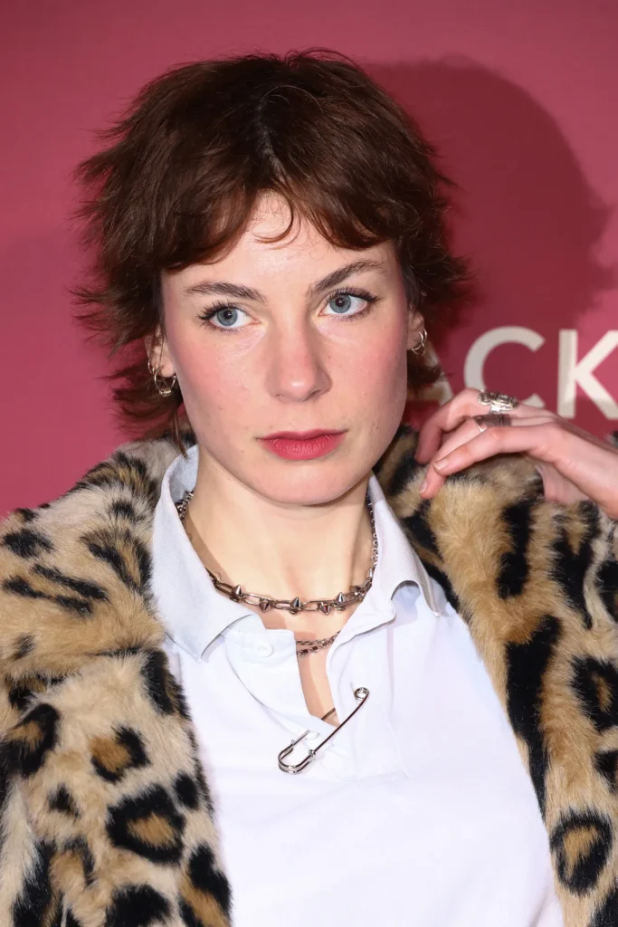 10 Stunning Pixie Haircuts for Black Women 4 A person wears a leopard print coat over a collared shirt, adorned with layered metal necklaces and rings, against a pink backdrop.