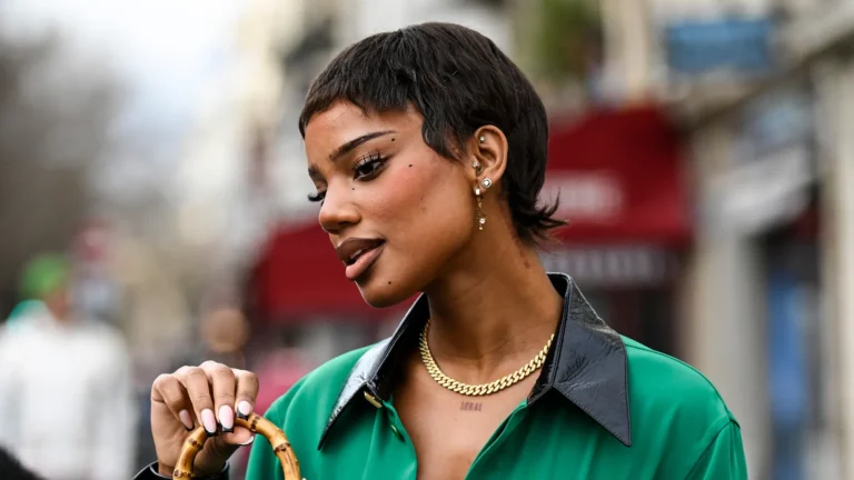 A fashionably dressed person in a green shirt with a black collar holds a bamboo handle bag, showcasing jewelry and urban style.
