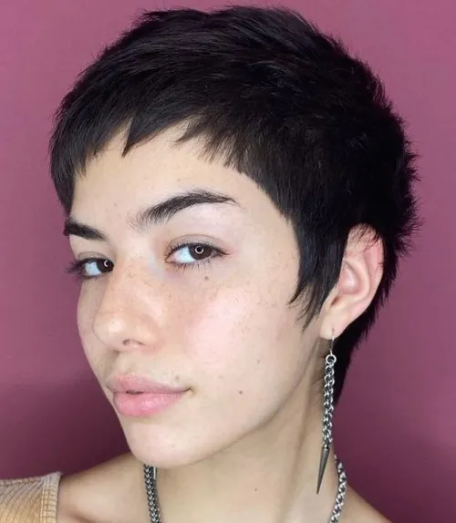 A person with short, textured hair wears a stylish earring, posed against a pink background, showcasing a modern and fashionable look.