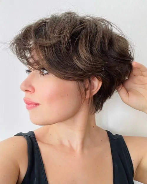 A person with short, tousled brown hair poses with one hand touching the back of their head, against a white background.